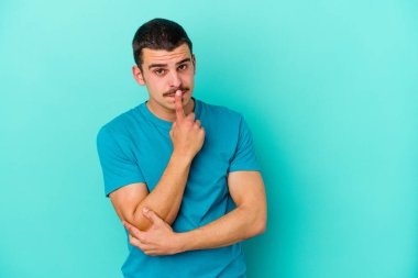 Mavi arka planda izole edilmiş genç beyaz adam alaycı bir ifadeyle kameraya bakarken mutsuz oluyor..