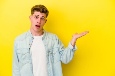 Sarı arka planda izole edilmiş genç beyaz adam, avuç içi fotokopi alanını elinde tutmaktan etkilendi..