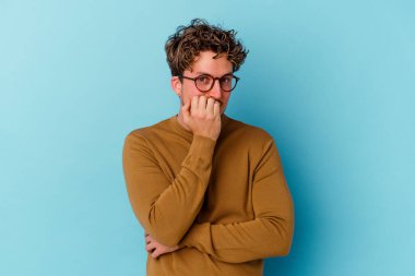 Mavi arka planda izole edilmiş, tırnak yiyen, gergin ve endişeli gözlüklü genç bir beyaz adam..