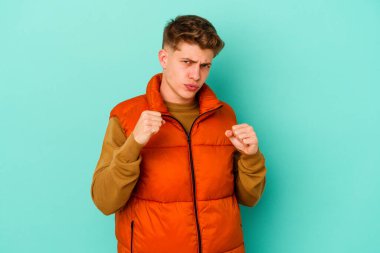 Mavi arka planda izole edilmiş genç beyaz adam yumruğunu kameraya gösteriyor, agresif yüz ifadesi var..