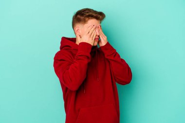 Mavi arka planda izole edilmiş genç beyaz adam elleriyle gözlerini kapatmaktan korkuyor..