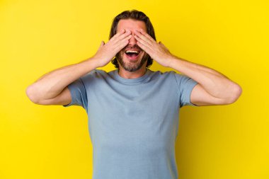 Sarı arka planda izole edilmiş orta yaşlı beyaz bir adam gözlerini elleriyle kapatıyor, geniş bir gülümsemeyle bir sürpriz bekliyor..