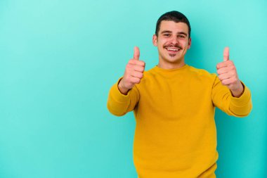 Mavi arka planda izole edilmiş genç beyaz adam gülümsüyor ve başparmağını kaldırıyor.