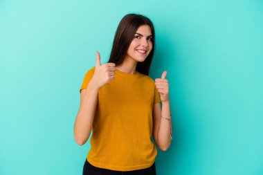 Kafkasyalı genç bir kadın mavi arka planda izole edilmiş. Baş parmağını kaldırıyor, gülümsüyor ve kendinden emin..