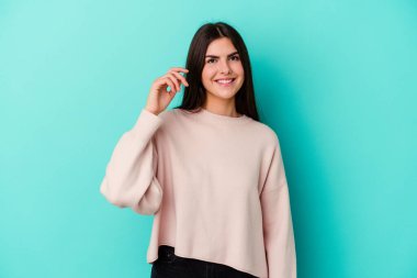 Mavi arka planda izole edilmiş genç beyaz kadın bir şeye gülüyor, elleriyle ağzını kapatıyor..