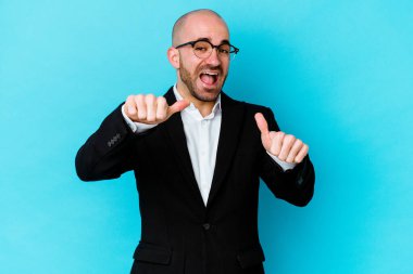 Genç, beyaz, kel bir adam mavi arka planda izole edilmiş. Baş parmağını kaldırıyor, gülümsüyor ve kendinden emin..