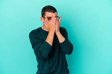 Mavi arka planda izole edilmiş genç beyaz adam kameraya parmaklarıyla göz kırpıyor, yüzünü kapatmaktan utanıyor..