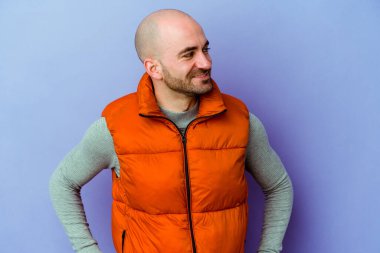 Mor arka planda izole genç beyaz kel adam mutlu bir şekilde gülüyor ve ellerini karnının üstünde tutarak eğleniyor..