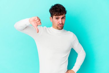 Mavi arka planda izole genç beyaz adam baş parmağını aşağı gösteriyor, hayal kırıklığı konsepti.