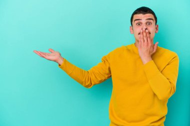 Mavi arka planda izole edilmiş genç beyaz adam, avuç içi fotokopi alanını elinde tutmaktan etkilenmiş..