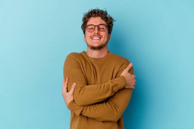 Mavi arka plan kucaklamalarında izole edilmiş gözlük takan genç beyaz adam, kaygısız ve mutlu gülümsüyor..