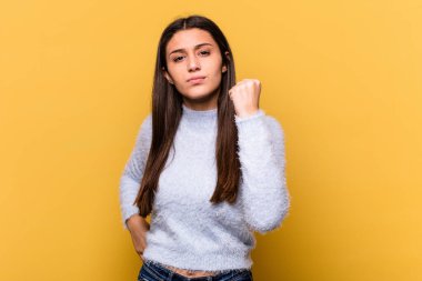 Sarı arka planda izole edilmiş genç Hintli kadın kameraya yumruk gösteriyor, agresif yüz ifadesi..