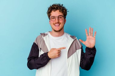Mavi arka planda izole edilmiş gözlüklü genç beyaz adam gülümseyerek beş numarayı parmaklarıyla gösteriyor..