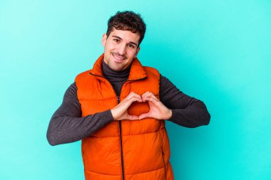 Mavi arka planda izole edilmiş genç beyaz adam gülümsüyor ve elleriyle kalp şeklini gösteriyor..