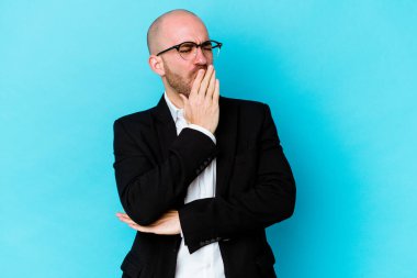 Genç, beyaz, kel bir adam mavi arka planda izole edilmiş esniyor. Ağzı eliyle kapatan yorgun bir hareket sergiliyor..