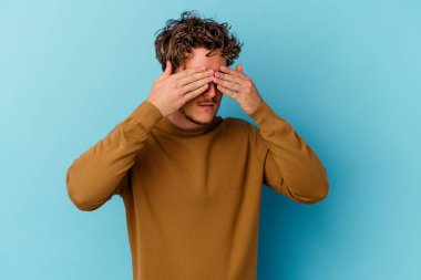 Mavi arka planda gözlük takan genç beyaz adam elleriyle gözlerini kapatmaktan korkuyor..