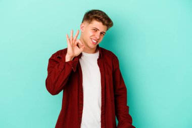 Kafkasyalı genç bir adam mavi arka planda izole edilmiş bir şekilde göz açıp kapayana kadar iyi bir el hareketi yapıyor..