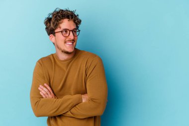 Mavi arka planda izole edilmiş gözlük takan genç beyaz adam kollarını kavuşturarak gülümsüyor..