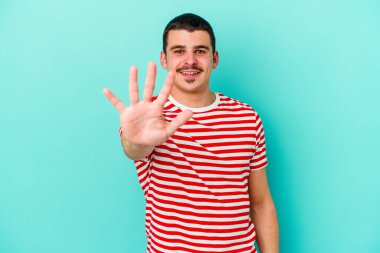 Mavi arka planda izole edilmiş genç beyaz adam gülümseyerek beş numarayı parmaklarıyla gösteriyor..