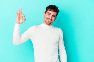 Kafkasyalı genç bir adam mavi arka planda izole edilmiş bir şekilde göz açıp kapayana kadar iyi bir el hareketi yapıyor..