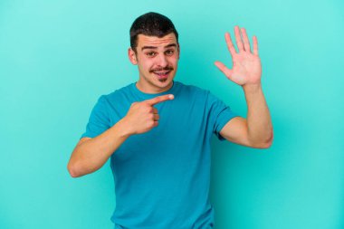Mavi arka planda izole edilmiş genç beyaz adam gülümseyerek beş numarayı parmaklarıyla gösteriyor..