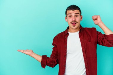 Mavi arka planda izole edilmiş genç beyaz bir adam avucunun içinde fotokopi alanı tutuyor, el üstünde tutuyor. Şaşırmış ve mutlu..