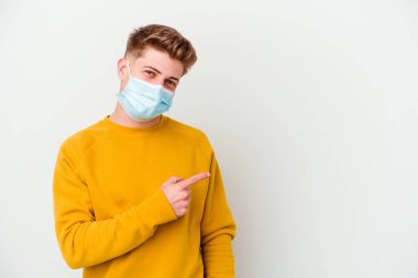 Koronavirüs maskesi takan genç adam beyaz arka planda izole edilmiş gülümseyen ve işaret eden boş alanda bir şey gösteriyor..