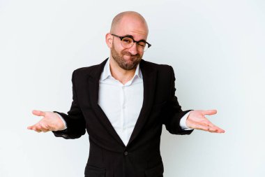 Genç, beyaz, kel bir adam mavi arka planda izole edilmiş. Sorgulama sırasında omuz silkiyor ve şüphe ediyor..