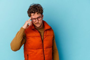 Gözlüklü genç beyaz adam mavi arka planda izole edilmiş. Şakağı parmağıyla işaret ediyor. Düşünüyor, bir göreve odaklanıyor..