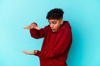 Genç Afro-Amerikan Kıvırcık Adam mavi üzerine izole edilmiş. Şaşırmış ve şaşırmış bir şekilde ellerinin arasında bir kopya alanı tutuyor..