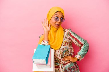 Genç Müslüman bir kadın pembe arka planda izole edilmiş giysiler alıyor. Dedikoduları dinlemeye çalışıyor..