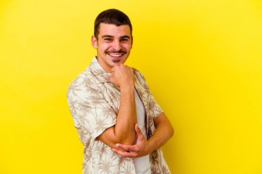 Sarı arka planda izole edilmiş, mutlu ve kendinden emin gülümseyen, elleriyle çeneye dokunan genç beyaz bir adam..