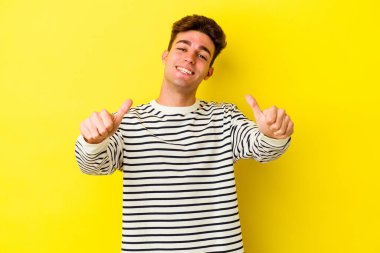 Sarı arka planda izole edilmiş genç beyaz adam iki parmağını da kaldırıyor, gülümsüyor ve kendinden emin..