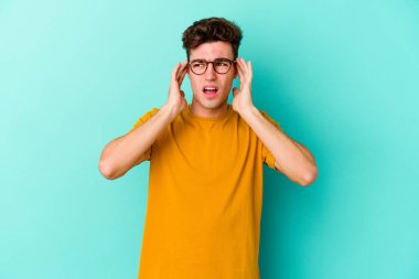 Genç beyaz adam mavi arka planda izole edilmiş parmaklarıyla kulaklarını kapatıyor, gürültülü bir ortam tarafından stresli ve umutsuz..