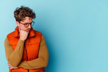 Mavi arka planda gözlük takan genç beyaz adam şüpheli ve şüpheci bir ifadeyle yana bakıyor..