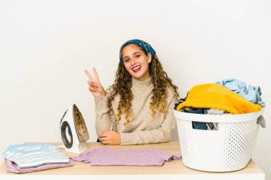 Giysileri ütüleyen beyaz bir kadın neşeli ve tasasız bir şekilde parmaklarıyla barış sembolü gösteriyor..