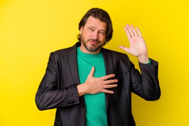 Orta yaşlı beyaz bir adam sarı arka planda izole edilmiş yemin ediyor, elini göğsüne koyuyor..