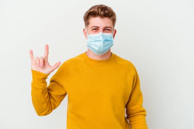 Koronavirüs maskesi takan genç bir adam beyaz arka planda izole edilmiş bir şekilde boynuz hareketi bir devrim konsepti olarak gösteriyor..