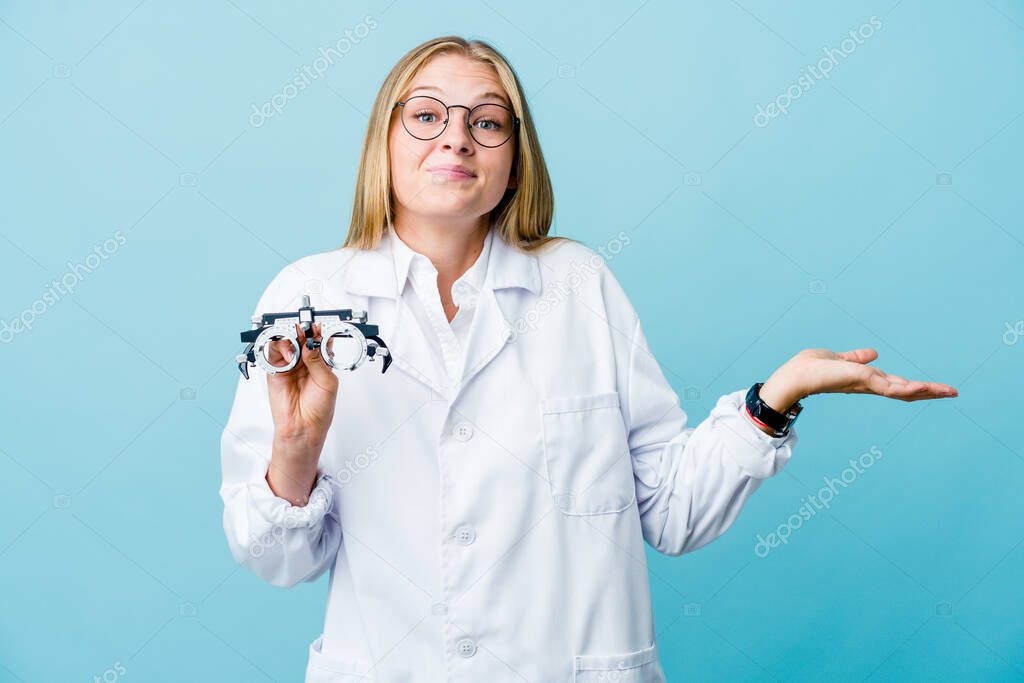 Young russian optometrist woman on blue doubting and shrugging shoulders in questioning gesture.