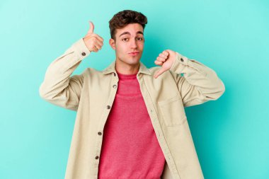 Kafkasyalı genç adam mavi arka planda izole edilmiş, baş parmağını kaldırıyor, zor seçim konsepti.