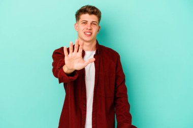 Mavi arka planda izole edilmiş genç beyaz adam gülümseyerek beş numarayı parmaklarıyla gösteriyor..