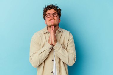 Mavi arka planda izole edilmiş gözlük takan genç, beyaz bir adam. Ağız kenarında el ele dua ediyor. Kendine güveniyor..