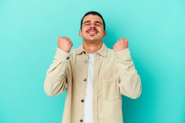 Mavi arka planda izole edilmiş genç beyaz bir adam zaferi, tutkuyu ve coşkuyu kutluyor, mutlu bir ifade..