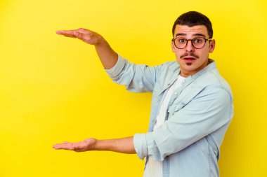 Sarı arka planda izole genç beyaz adam şok ve hayret içinde elleri arasında bir kopya alanı tutuyor.