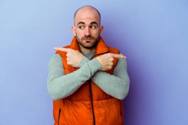 Mor arka plan noktalarında izole genç beyaz adam, iki seçenek arasında seçim yapmaya çalışıyor.