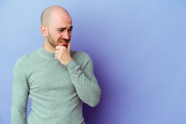 Kafkasyalı kel adam mor arka planda izole edilmiş kafasının arkasına dokunuyor, düşünüyor ve bir seçim yapıyor..