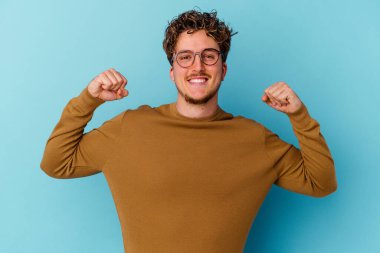 Mavi arka planda gözlük takan genç beyaz adam kadınsı gücün sembolü olan kollarıyla güçlü bir hareket sergiliyor.