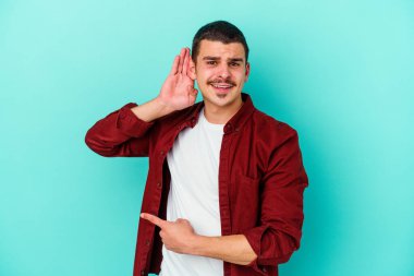 Mavi arka planda izole edilmiş genç beyaz adam bir dedikoduyu dinlemeye çalışıyor..