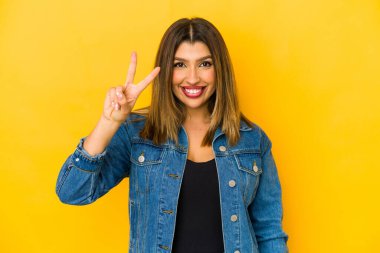 Sarı arka planda izole edilmiş genç Hintli kadın neşeli ve kaygısız parmakları olan bir barış sembolü gösteriyor..