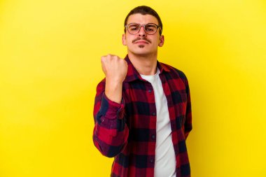 Sarı arka planda izole edilmiş genç beyaz adam kameraya yumruğunu gösteriyor, agresif yüz ifadesi..